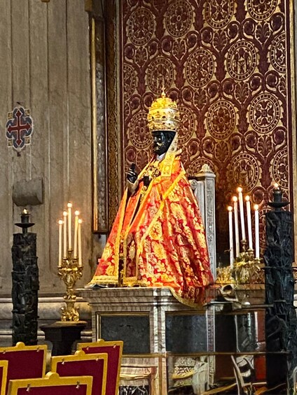 A statue in a richly decorated church interior with ornate wall patterns, lit candles, and a cross on the wall. 3