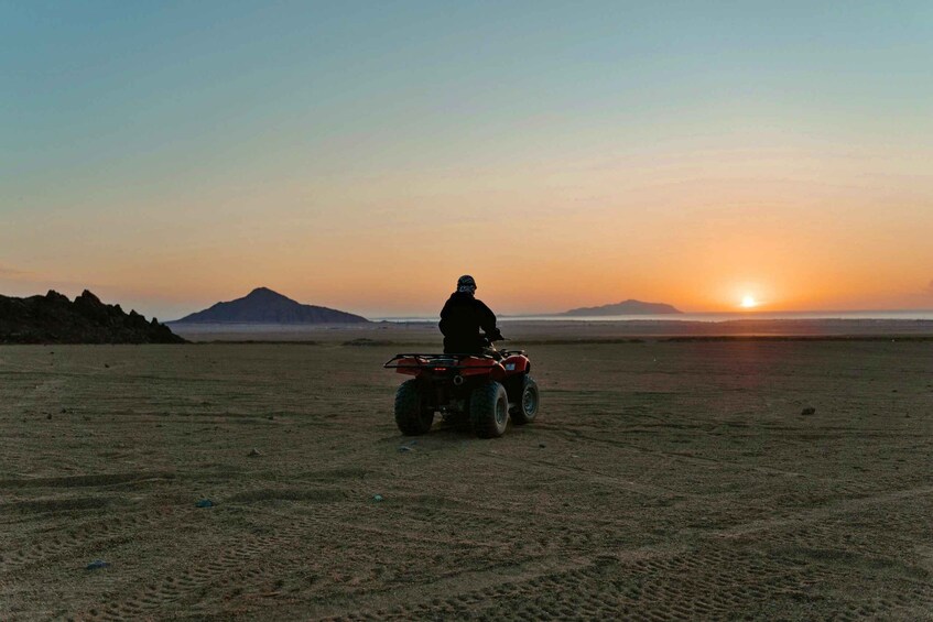 A person riding a red ATV on a flat desert landscape with mountains in the distance and a sunset. 3