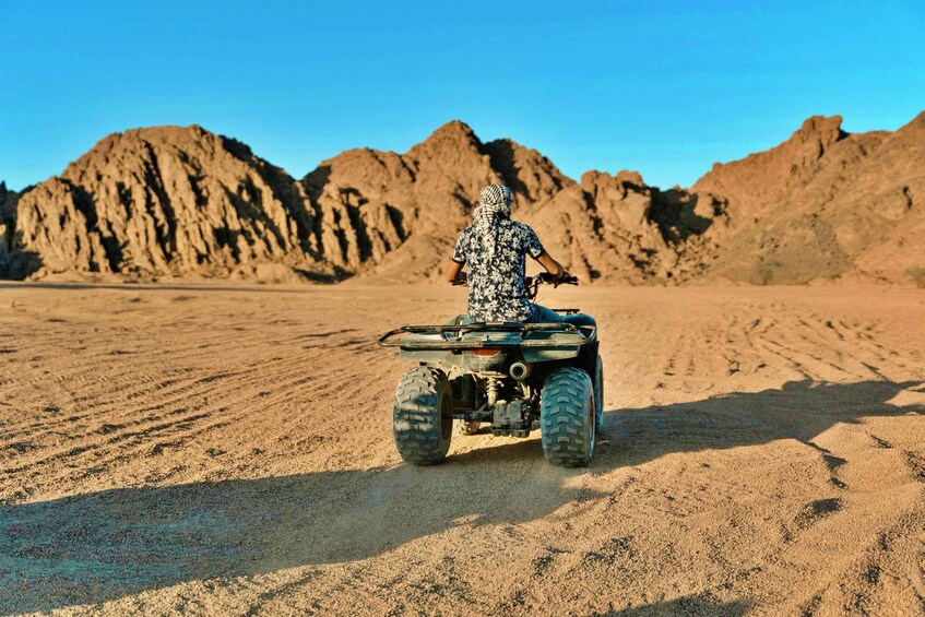 A person riding an ATV in a desert with rocky mountains in the background. 1