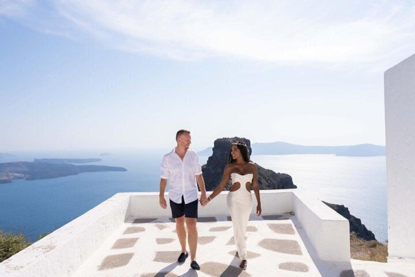 Un couple se tenant la main sur une terrasse aux murs blancs, d’où l’on aperçoit une étendue d’eau et des reliefs lointains. 1