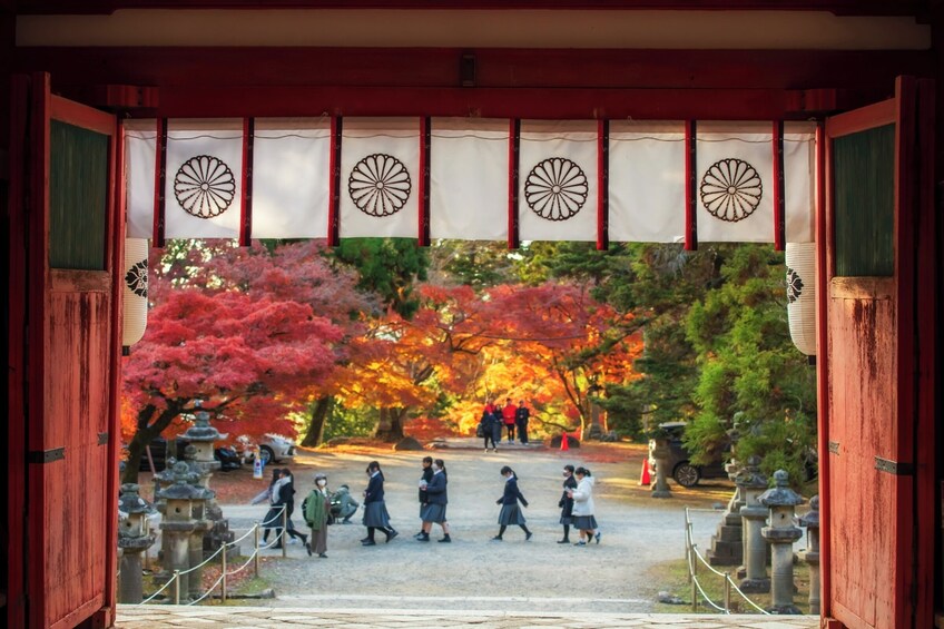 En traditionel japansk port med røde søjler og dekorative mønstre, der fører til en park med træer i efterårsfarver og stenlanterner. 2