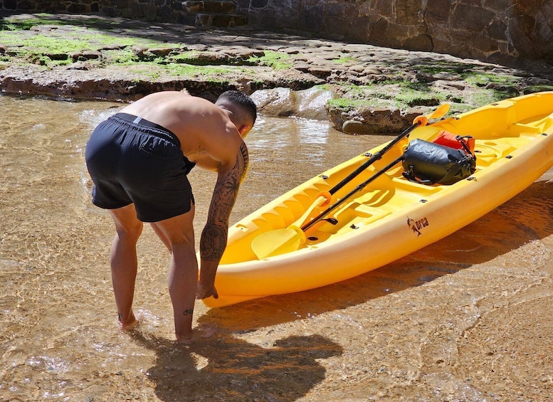 A person in black shorts preparing a yellow kayak with paddles and a black bag on a rocky shore. 5