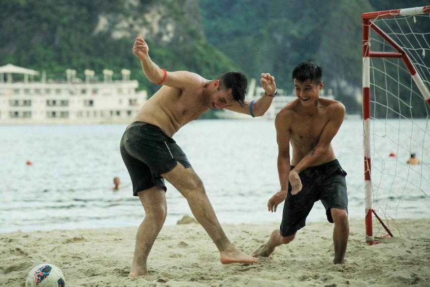 Two people playing soccer on a beach with a goalpost in the background. 1