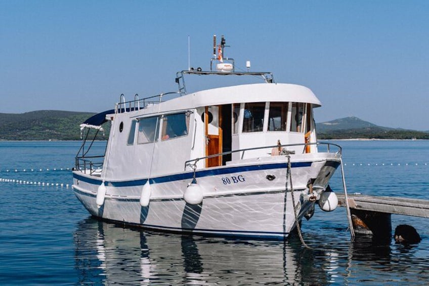 Ein weißes Boot mit blauen Streifen und der Aufschrift 80 BG, liegt an einem Steg. Im Hintergrund ist ruhiges Wasser und eine hügelige Landschaft zu sehen. 3