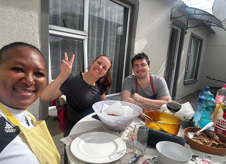 Three people seated at an outdoor table with food and drinks, a clear sky, and a building in the background. 5
