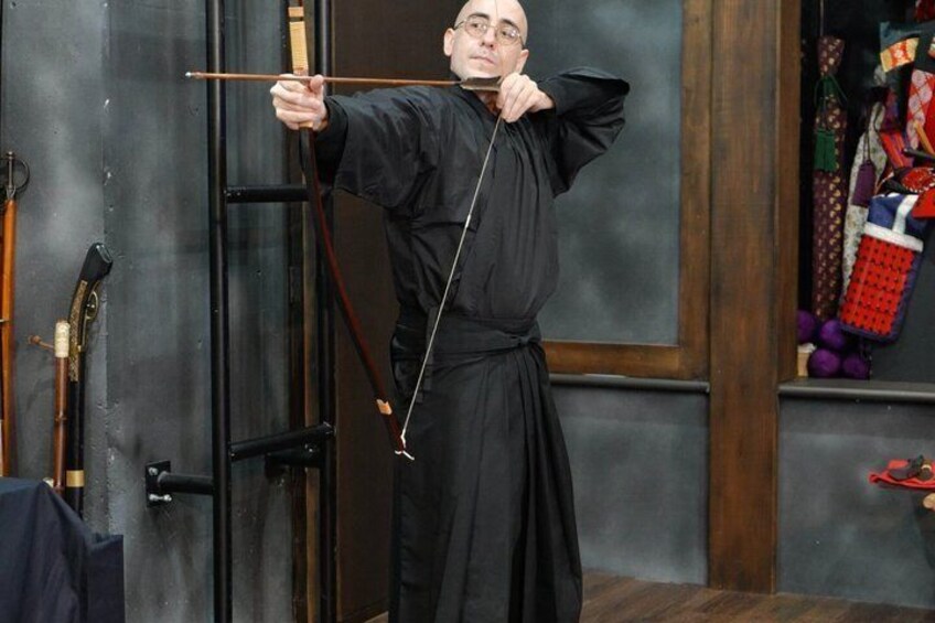 Una persona vestita di nero che scocca frecce con un arco, in una stanza con pavimento in legno e parete nera. 2