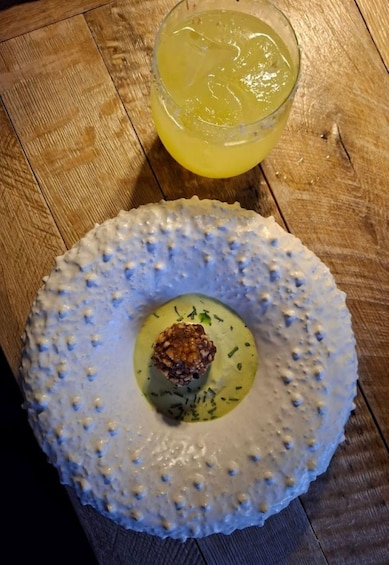 A round, textured plate with a central garnish, accompanied by a glass of yellow beverage on a wooden table. 4