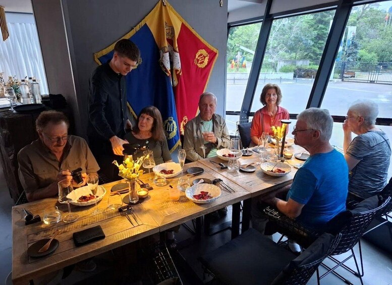 A group of people seated around a wooden table with plates of food, glasses of wine, and a flag in the background. 3