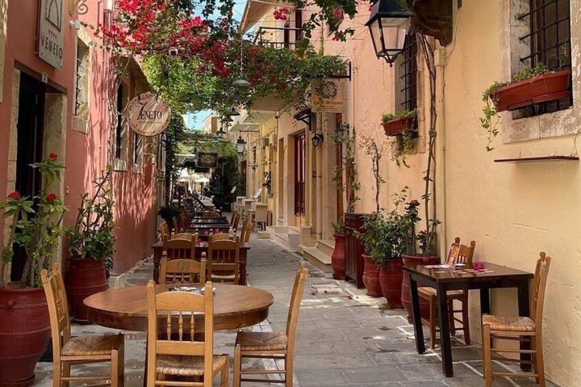 A narrow street with tables and chairs, potted plants, and hanging plants. 3