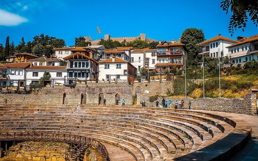 An ancient amphitheater with stone seating, surrounded by traditional buildings and a hilltop fortress. 4