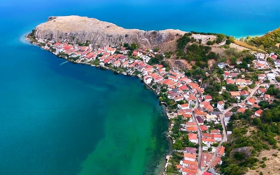 An aerial view of a coastal town with red-roofed houses, a clear blue sea, and a mountainous backdrop. 1