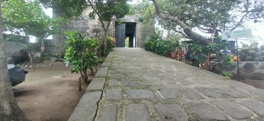 A stone pathway leading to a dark doorway, surrounded by trees and a small structure with a sign. 5