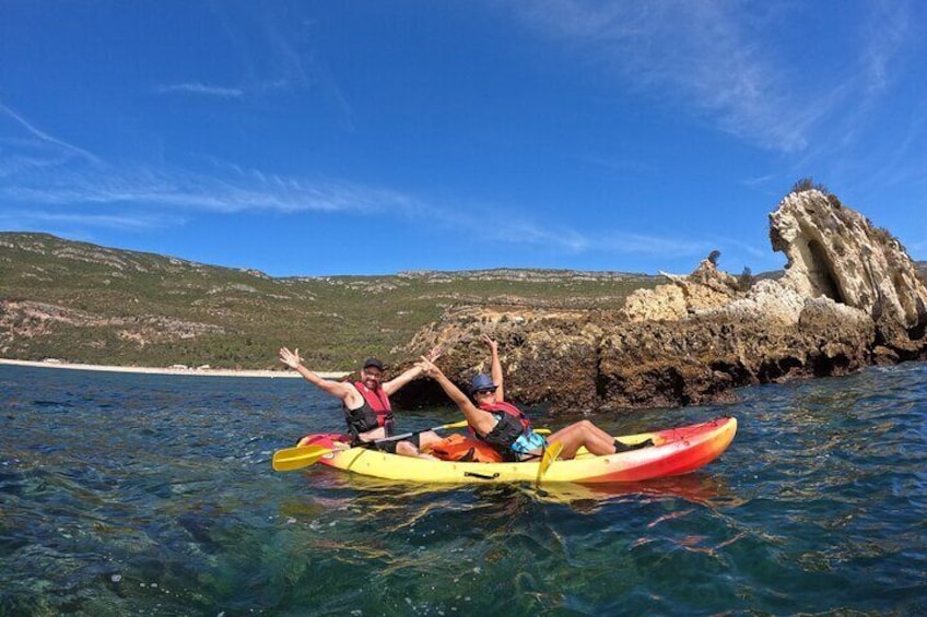 Two people kayaking on clear water with a rocky coastline and cliffs in the background. 1