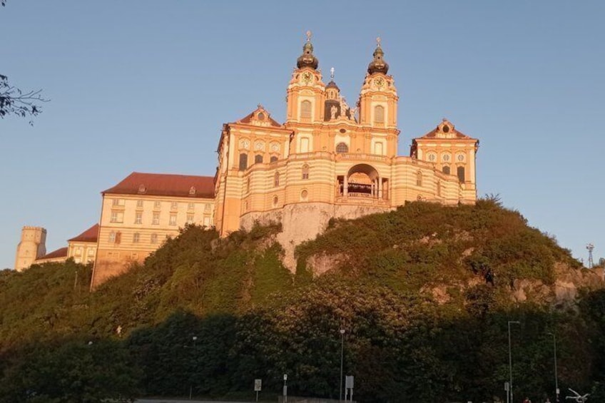 A large, historic building with multiple towers, situated on a hill with dense greenery. 1