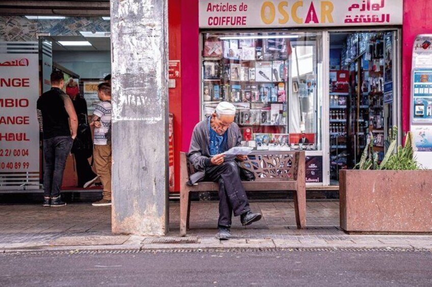 A man sitting on a bench outside a store with a sign that reads 'Articles de COIFFURE OSCAR.' 3