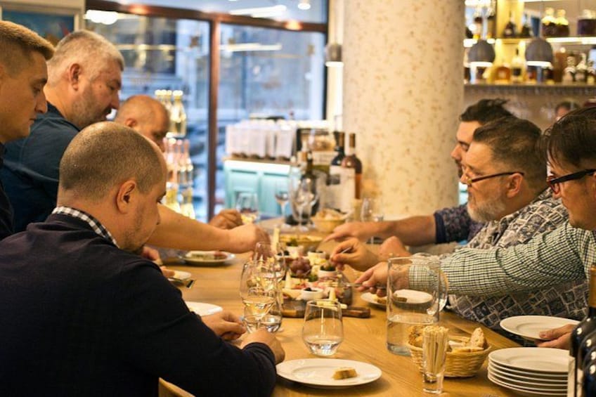 En grupp människor som äter middag på en restaurang, vid ett långt bord med vinglas och tallrikar fyllda med mat. 2