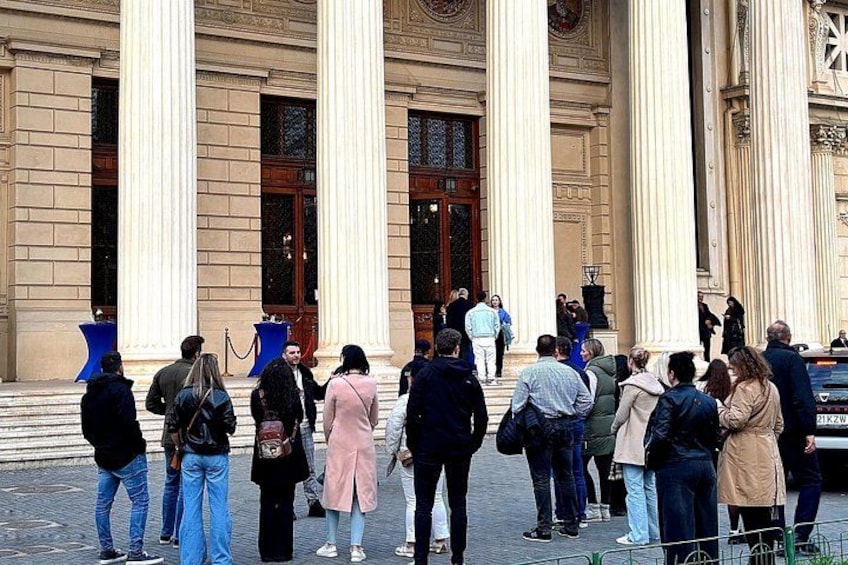 Un gruppo di persone in piedi di fronte a un edificio con colonne imponenti e un orologio. 3