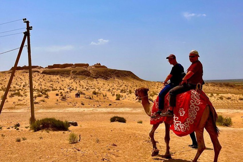 Two people riding a camel in a desert landscape with a power line structure and distant rocky formations. 5