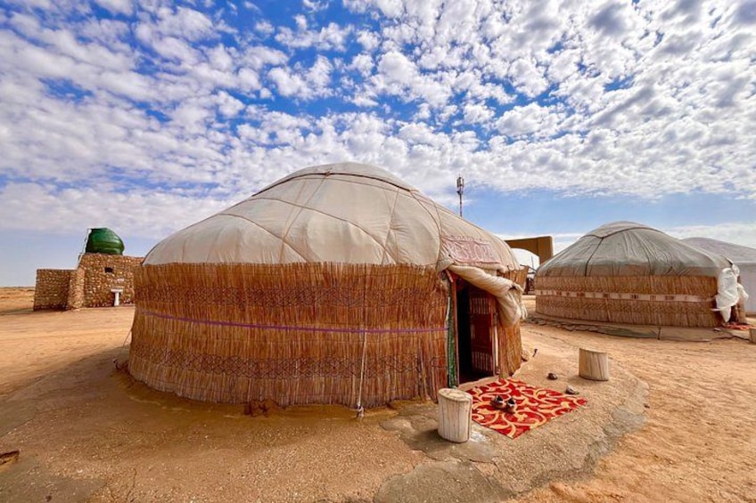 A traditional yurt with a thatched roof, a stone wall, and a red patterned rug on the ground. 1