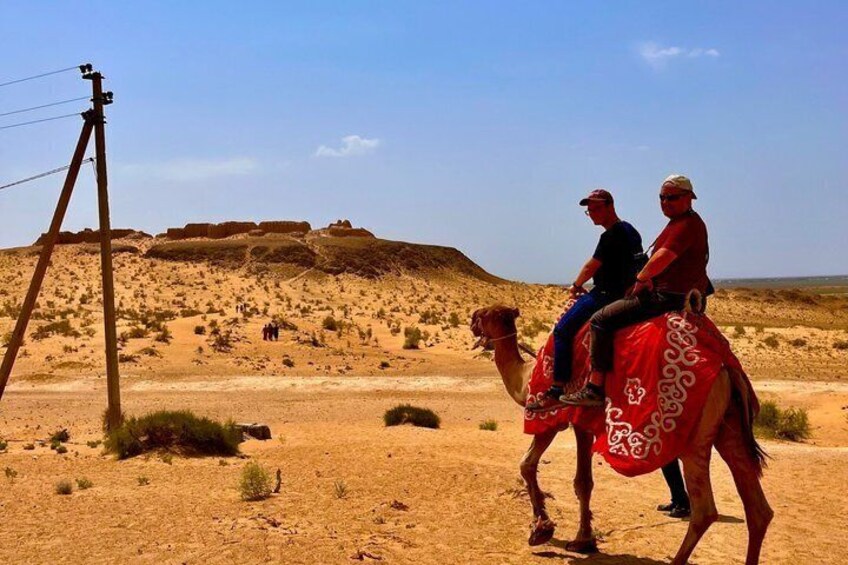Two people riding a camel in a desert landscape with a power line in the foreground. 5