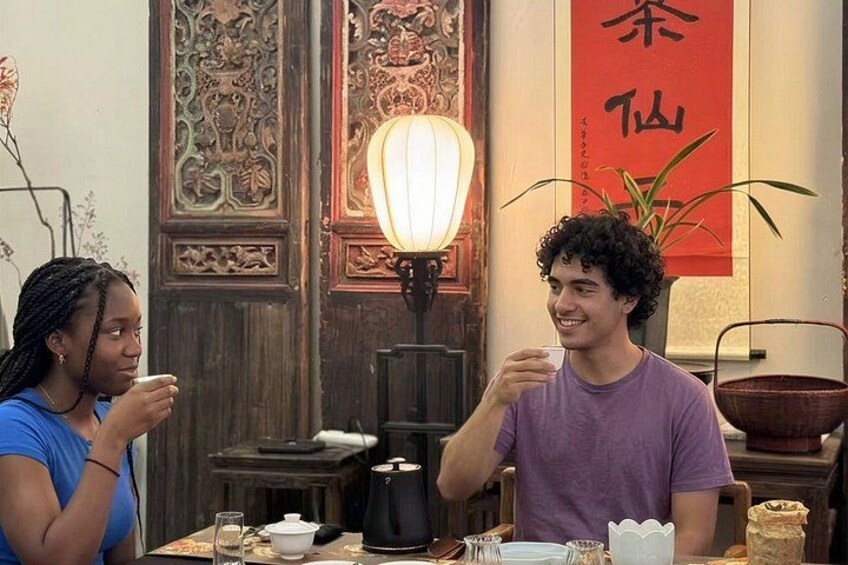 Zwei Personen sitzen an einem Tisch mit einem traditionellen Tee-Set, einer Lampe und einem roten Banner mit chinesischen Schriftzeichen. 4
