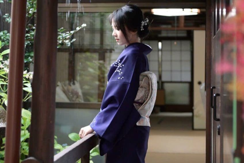 Una persona vestida con traje tradicional está de pie junto a una puerta de cristal con marco de madera, un banco de madera y vistas a un interior japonés tradicional. 1