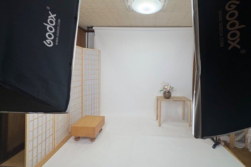 A minimalist indoor setting with a white floor, a small wooden stool, a table with a vase, and a black backdrop with the word 'Godox' on it. 2