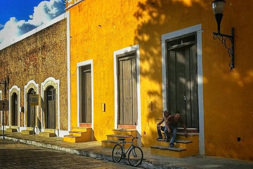 Un edificio amarillo con una bicicleta, una persona en los escalones y una farola. 1