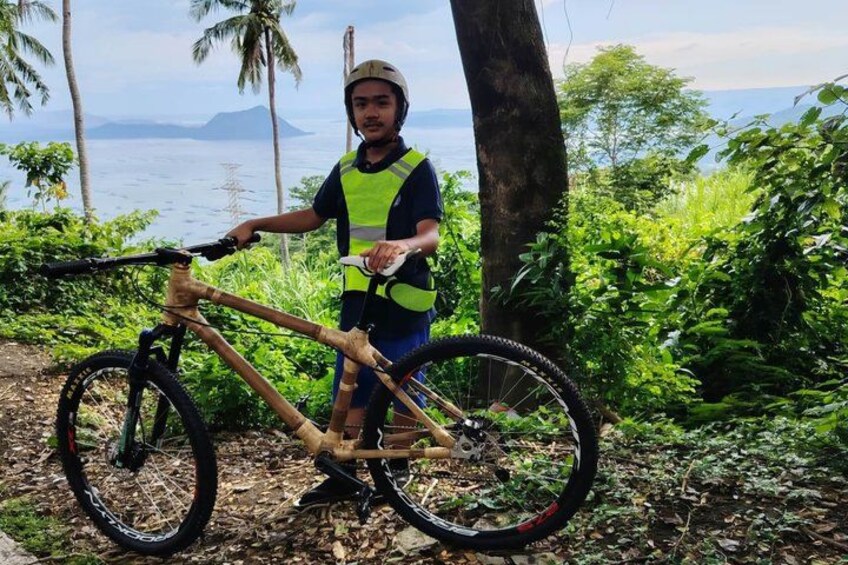 A person standing next to a mountain bike in a forested area with a view of the ocean and mountains in the background. 3