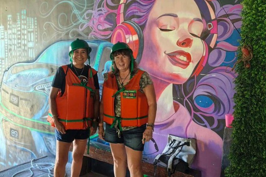 Deux personnes portant des gilets de sauvetage et des chapeaux, debout devant une fresque représentant un paysage urbain et des motifs abstraits. 4