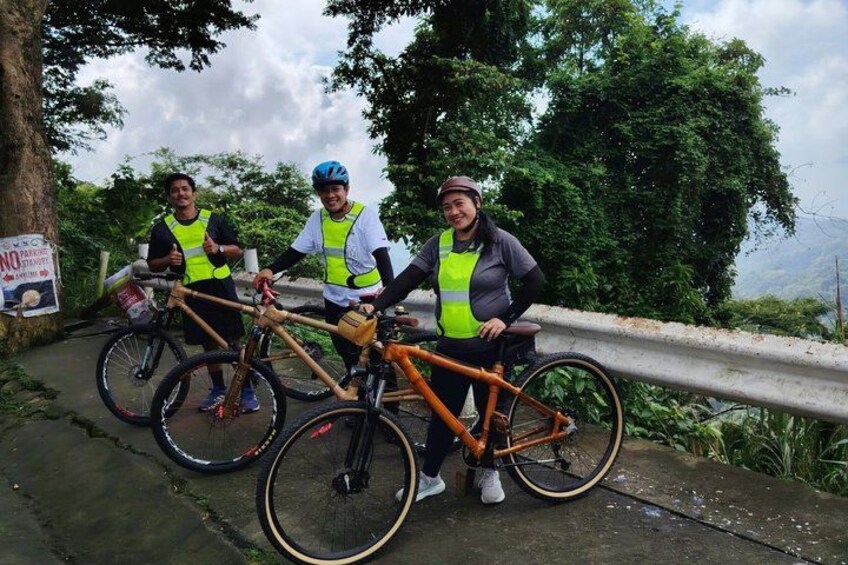 Three cyclists with bicycles on a paved path, surrounded by greenery and a mountainous landscape. 5