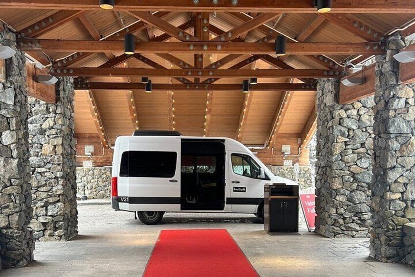 A white van parked under a wooden roof structure with stone walls, a red carpet, and a trash bin. 2