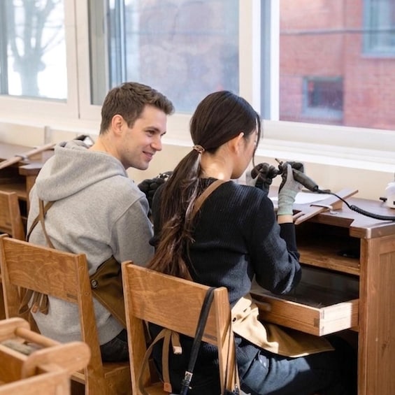 Two people seated at wooden desks in a classroom with books and papers. 1