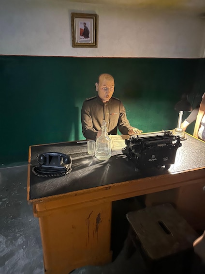 A man in a military uniform sitting at a desk with a telephone, a glass, and a typewriter. 5