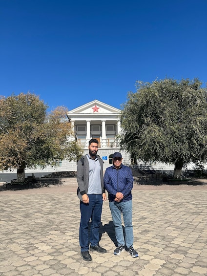 Two people standing in front of a building with a red star on the roof. 4