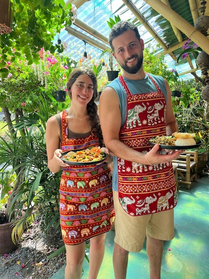 Two people in a greenhouse holding plates of food, surrounded by various plants and a bamboo structure. 2