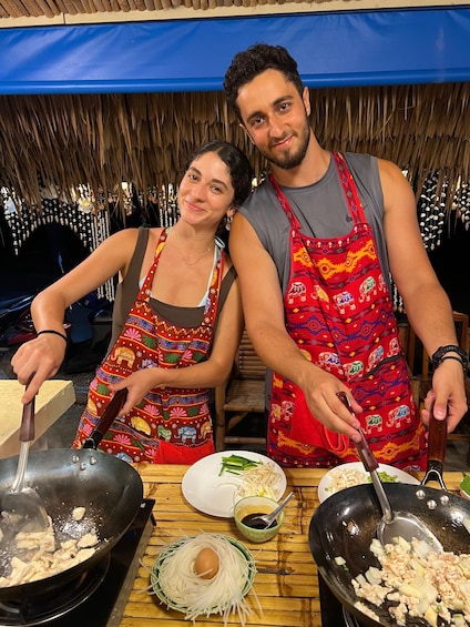 Two people cooking in a traditional setting with wooden tables and thatched roofs. 5