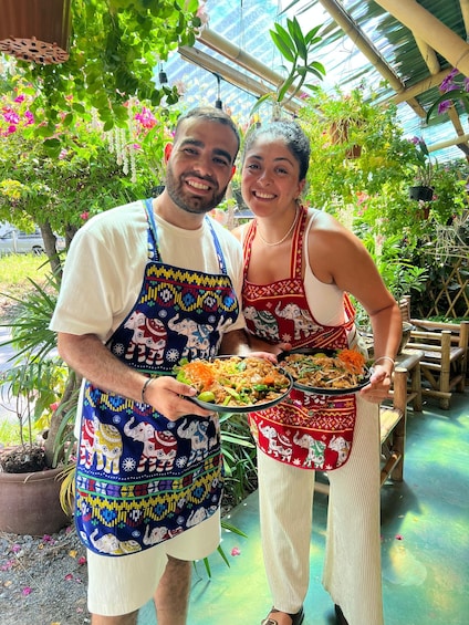 Two people holding plates of food in a greenhouse with various plants and a bamboo structure. 3