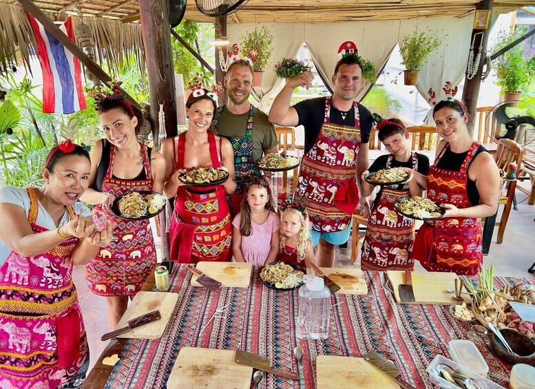A group of people in traditional attire holding plates of food, with a thatched roof and tropical plants in the background. 1