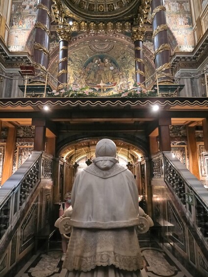 A statue of a robed figure in a grand hall with ornate columns and a detailed fresco above. 4