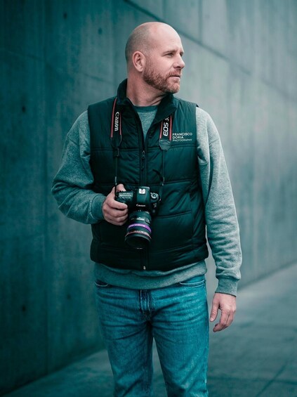 A person holding a Canon camera, wearing a green vest with 'Francisco Doria Photography' on it, standing in an urban setting. 3