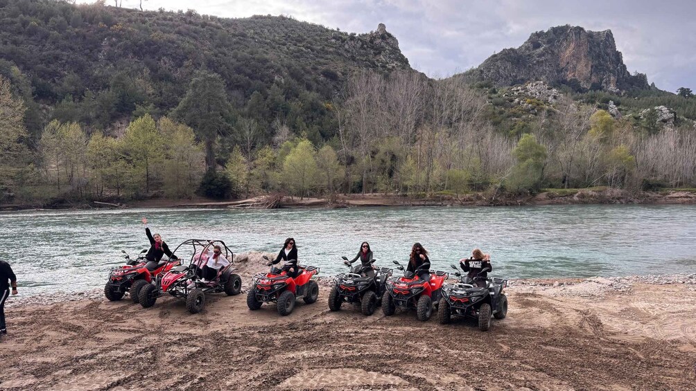 A group of off-road vehicles parked by a river with a mountainous backdrop. 4