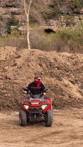 A person riding a red ATV on a dirt path with a pile of dirt in the background and a tree to the left. 2