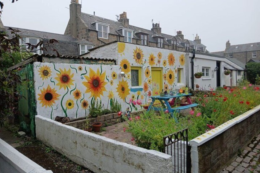 A house with a sunflower mural, a garden with red flowers, and a blue table. 1
