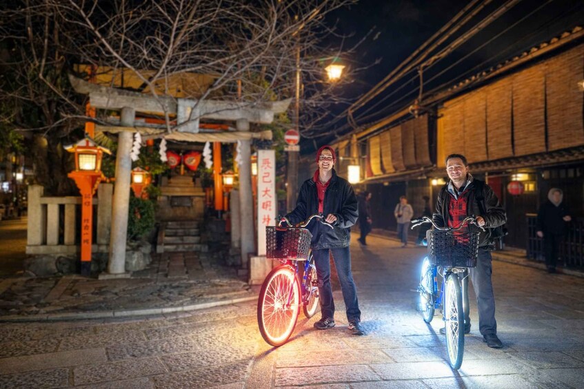 Two people on bicycles with baskets, a traditional Japanese torii gate, and a brick building in the background. 1