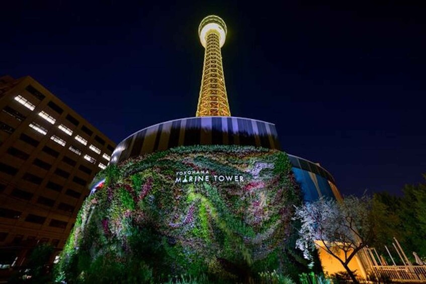 A night view of the Yokohama Marine Tower with a lit top and a digital display screen at its base. 3