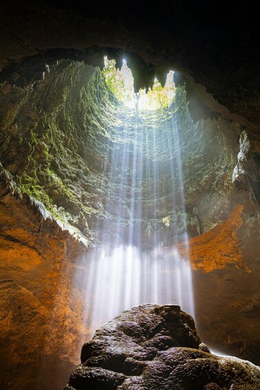 A cave with sunlight streaming through a hole, illuminating the interior and a large rock in the foreground. 5