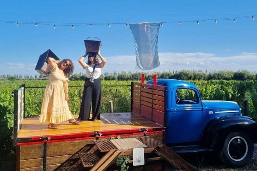 A blue vintage truck with a wooden platform, two people standing on it, and a vineyard in the background. 5