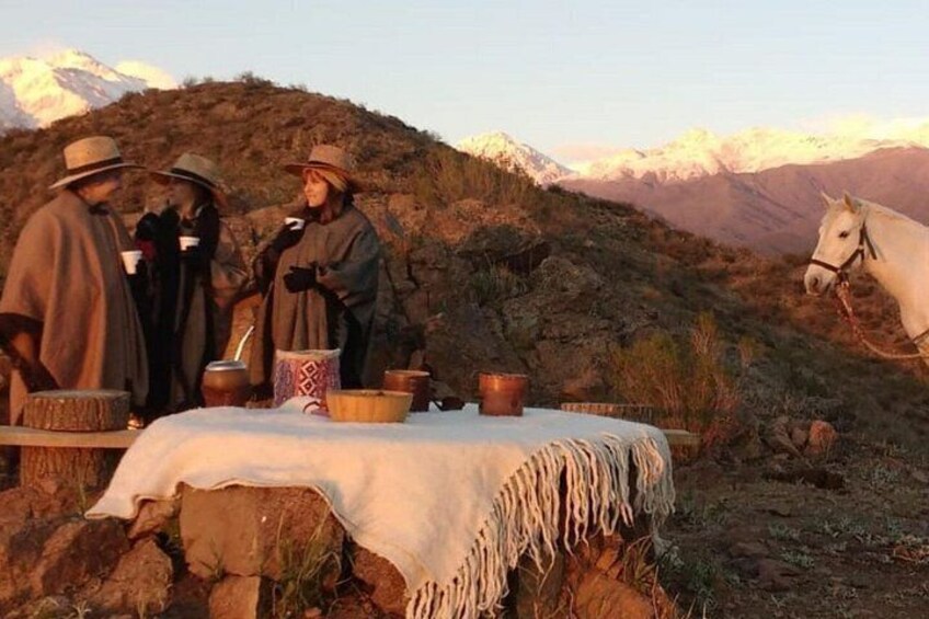 A group of people around a table with cooking utensils, a horse, and a mountainous landscape in the background. 1
