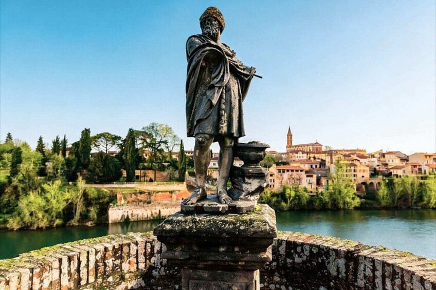 A statue on a stone platform by a river with buildings in the background. 4
